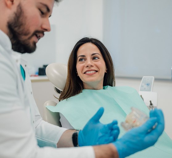 Woman in dental chair smiling at dentist holding model teeth discussing treatment